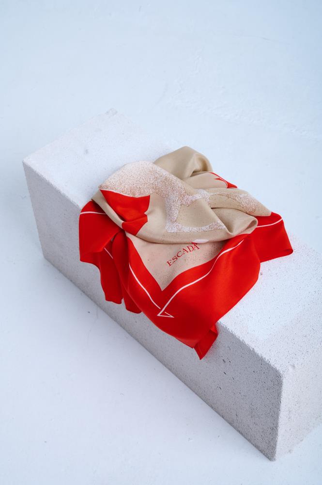 A luxury silk scarf in beige and vibrant red, featuring a minimalist graphic print and bold branding, elegantly draped over a white stone block in a studio setting.