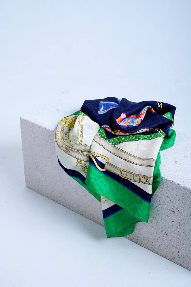A luxury silk scarf with a classic equestrian and nautical theme, featuring golden chains and leather belt prints on a navy blue, white, and emerald green background, draped over a white stone block.