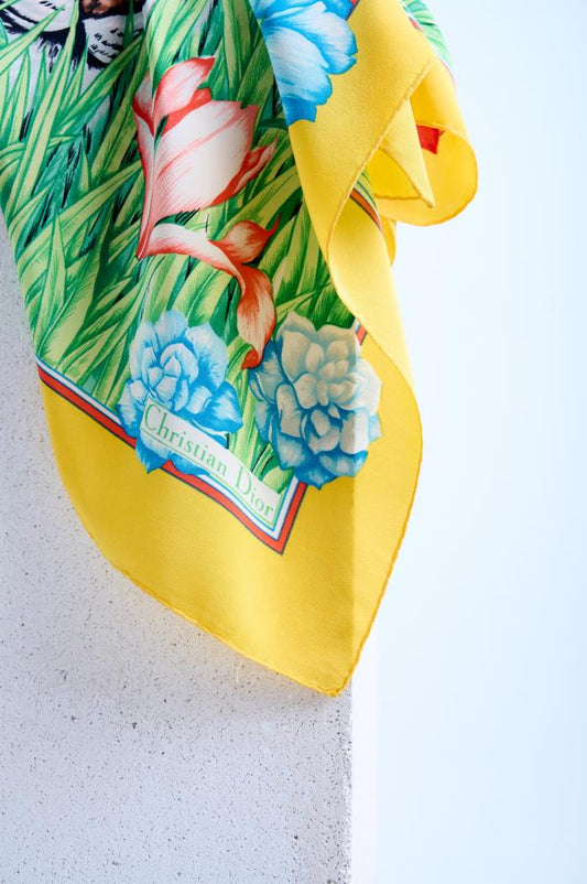 A yellow silk scarf with a vibrant floral and animal pattern, featuring a tiger in a tropical meadow, displayed on a wall.