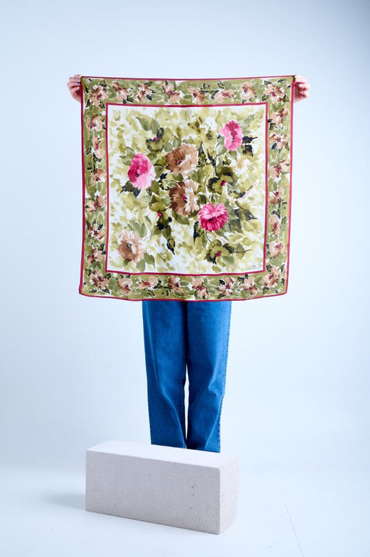 A person holding up a square silk scarf with a vintage floral pattern featuring large pink and brown carnations on a sage green and cream background, finished with a decorative floral border.