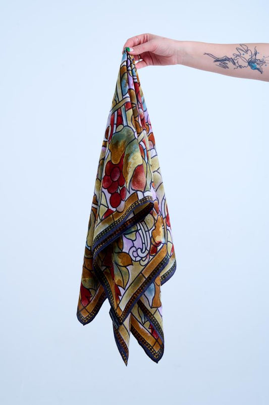 A hand with a tattoo holding up a colorful silk scarf with a stained-glass style print featuring grapes and autumn leaves in rich red, gold, and purple tones.