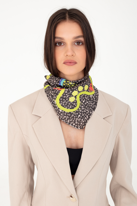 Fashion portrait of a woman wearing the "La Dolce Vita" dark brown patterned silk and viscose scarf featuring yellow lemon slice illustrations, styled with a light beige blazer against a plain white studio background