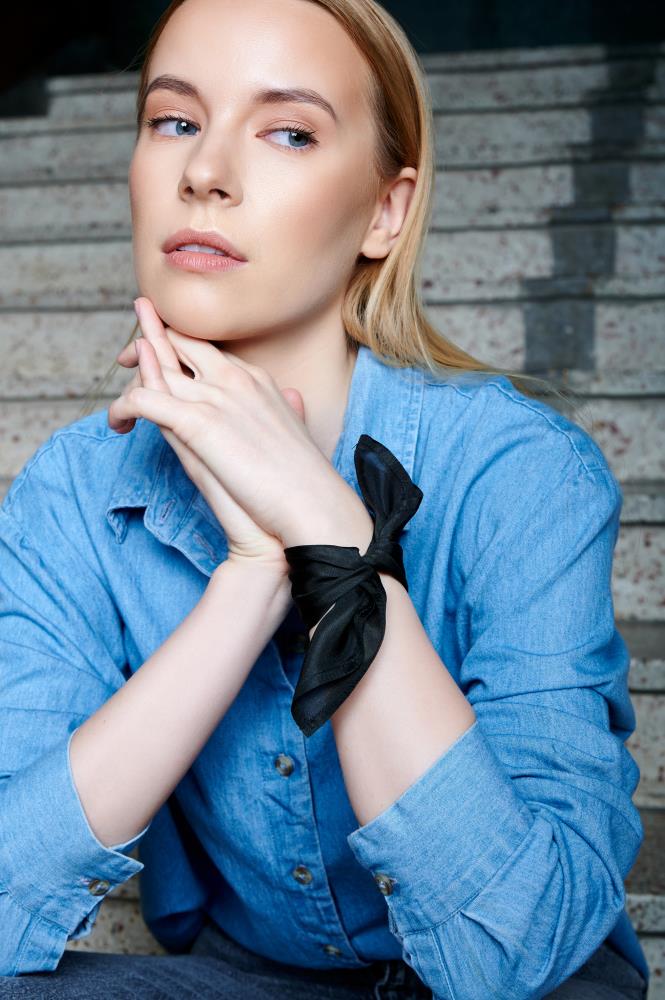 A thoughtful woman in a blue denim shirt sits on stone steps, hands clasped under her chin. She wears a dark moss green scarf on her wrist and gazes to the side.