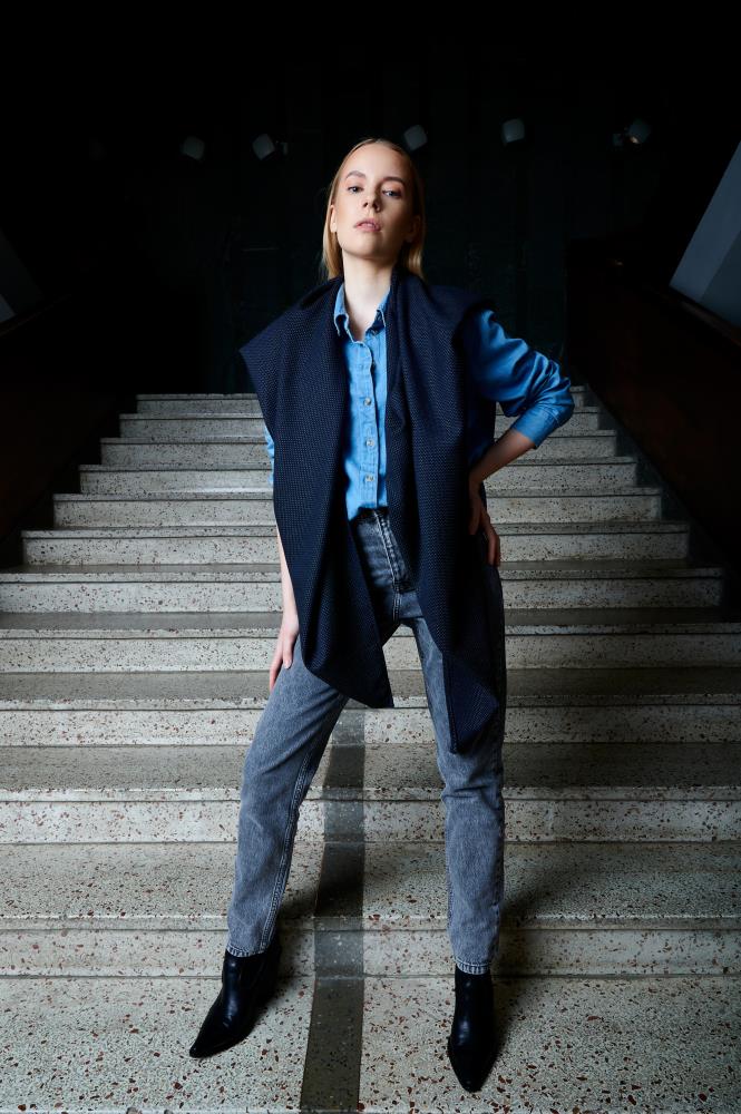A full-length photo of a woman posing on wide stone stairs in a dark interior. She is wearing a light blue denim shirt, grey jeans, and black pointed boots. A wide navy blue scarf is stylishly draped over her shoulders, falling freely in front to add a modern touch and volume to the outfit. The lighting is focused on the woman, highlighting the clothing details and creating a dramatic contrast with the dark background.