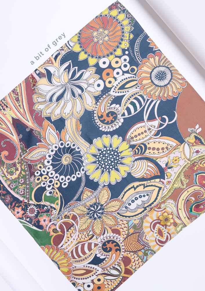 A  silk scarf featuring a playful and colorful bohemian pattern. The design combines oriental paisley motifs with large, stylized flower blossoms against a rich navy blue background. The color palette intertwines coral, yellow, green, and terracotta brown hues, subtly complemented by light grey elements. The text "a bit of grey" in the photo highlights this color accent and the characteristic elegance of the accessory.
