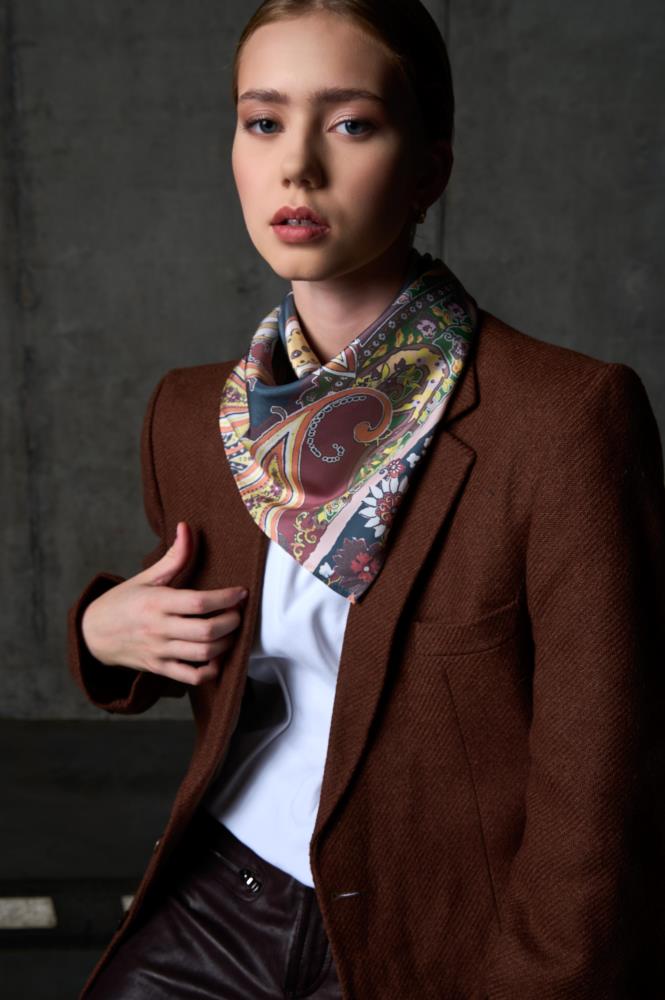 A portrait of a woman posing against a dark concrete wall in an industrial setting. She is wearing a brown wool blazer over a white t-shirt and dark leather trousers. A vibrant silk scarf with an intricate floral and paisley pattern in shades of burgundy, mustard, and teal is elegantly draped around her neck. The lighting creates a sophisticated mood, highlighting the contrast between the textured blazer and the smooth, patterned silk accessory.