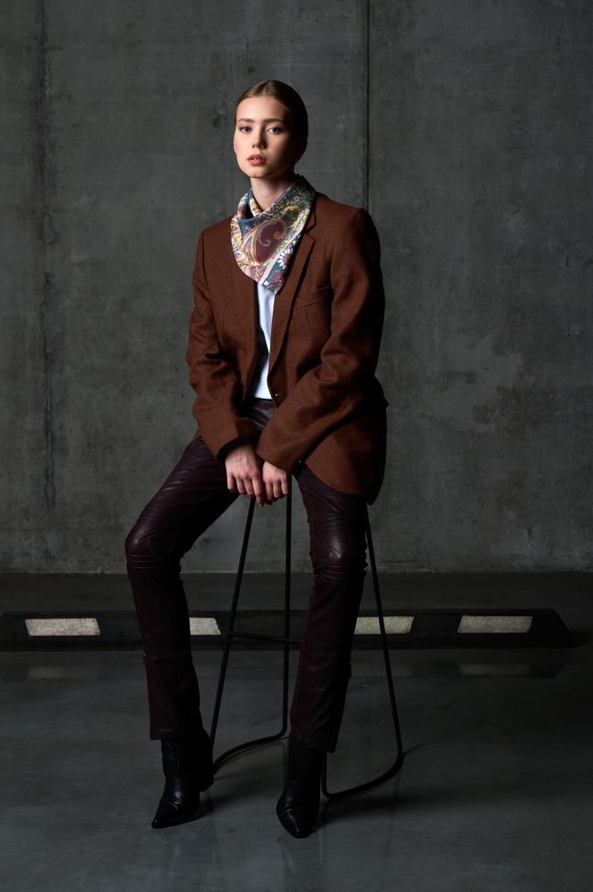 A full-length photo of a woman sitting on a high bar stool against a dark concrete wall in an industrial setting. She is wearing a brown wool blazer, dark burgundy leather trousers, and black pointed-toe boots. A colorful silk scarf with an intricate paisley and floral pattern is neatly tied around her neck. The minimalist lighting and raw background create a sophisticated, edgy aesthetic that emphasizes the textures and colors of the outfit.