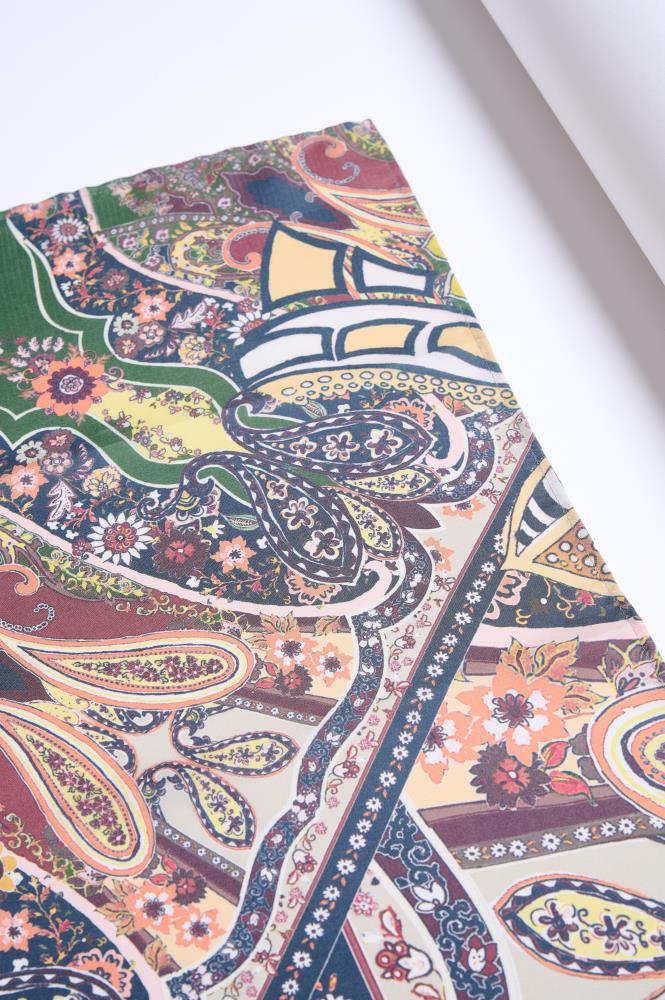 A top-down, close-up photo of a colorful silk scarf displayed on a white surface. The fabric is adorned with an intricate and dense pattern featuring traditional paisley motifs and various floral designs. The color palette is rich and diverse, including shades of forest green, burgundy, mustard yellow, and teal, accented with small white flower borders. The lighting is bright and even, highlighting the detailed print and the smooth texture of the silk.