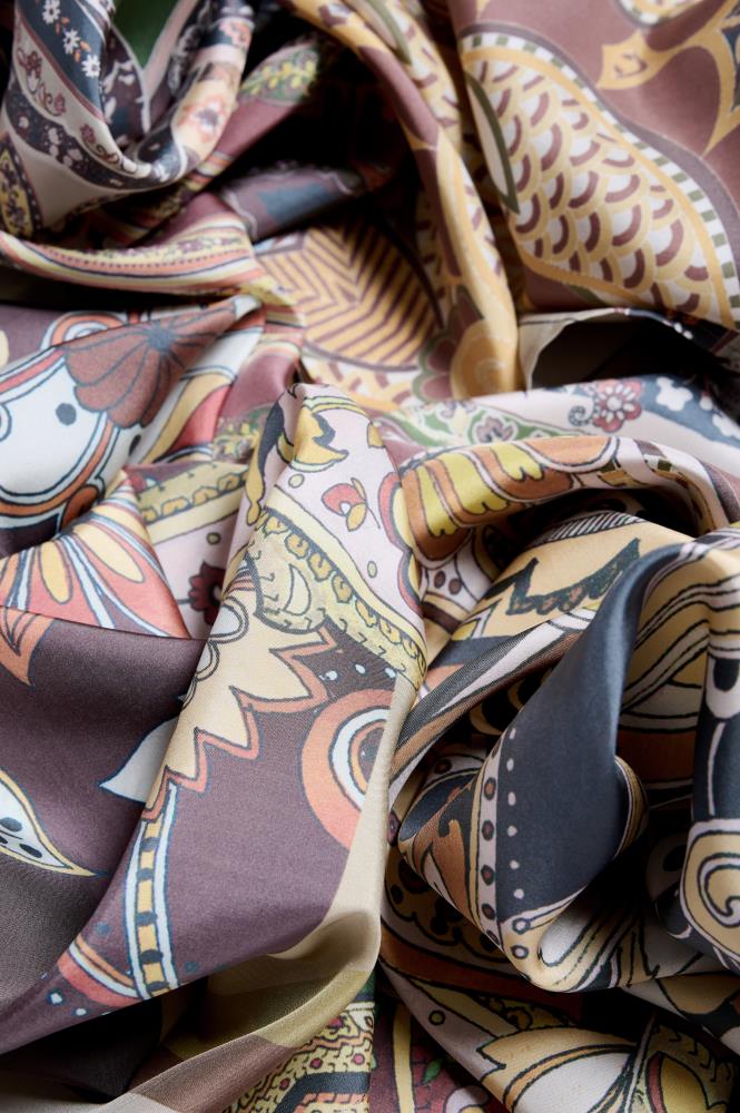 An extreme close-up photo of a luxurious silk scarf with a complex, patterned print. The fabric is artfully crumpled into numerous rich folds and creases, creating a dynamic composition of textures and shadows. The intricate print features varied traditional paisley and abstract floral motifs in an earthy color palette of burgundy, mustard yellow, teal, and cream, with black outlines. Soft, directional light highlights the detailed patterns and the smooth sheen of the silk material.