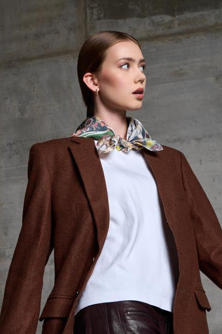 A low-angle portrait of a woman looking away from the camera, posed against a textured grey concrete wall. She is wearing a structured brown wool blazer over a crisp white t-shirt, paired with dark burgundy leather trousers. A vibrant silk scarf with an intricate, colorful pattern is elegantly knotted around her neck. The lighting highlights the clean lines of her outfit and the sophisticated contrast between the industrial background and the refined silk accessory.