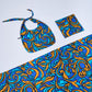 Flat lay of a 3-piece cotton collection with a blue, cyan, and bronze baroque swirl pattern. Includes a folded blanket, a soft tote bag with knotted straps, and a matching scarf, beautifully arranged on a light gray background.