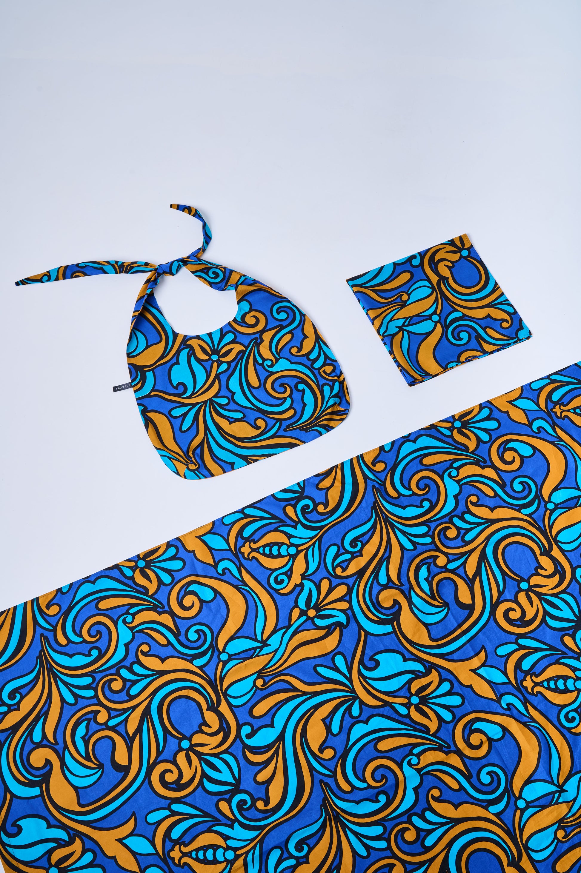 Flat lay of a 3-piece cotton collection with a blue, cyan, and bronze baroque swirl pattern. Includes a folded blanket, a soft tote bag with knotted straps, and a matching scarf, beautifully arranged on a light gray background.