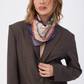 A woman with wavy blonde hair wearing the GUARDIAN Blue Graphite Silk Scarf tied neatly around her neck. The scarf features a deep graphite blue and charcoal border with a cream center and blue line art. She is styled in an oversized dark brown blazer, creating a sophisticated and modern look against a white background.