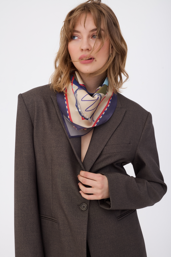 A woman with wavy blonde hair wearing the GUARDIAN Blue Graphite Silk Scarf tied neatly around her neck. The scarf features a deep graphite blue and charcoal border with a cream center and blue line art. She is styled in an oversized dark brown blazer, creating a sophisticated and modern look against a white background.
