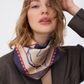 A close-up portrait of a woman with messy blonde hair, looking directly at the camera with her hand raised to her head. She is wearing the GUARDIAN Blue Graphite Silk Scarf tied loosely around her neck. The scarf's creamy center features a blue line-art terrier illustration, framed by a deep graphite blue border with a red and white dashed line. She is wearing a dark brown blazer against a bright white background