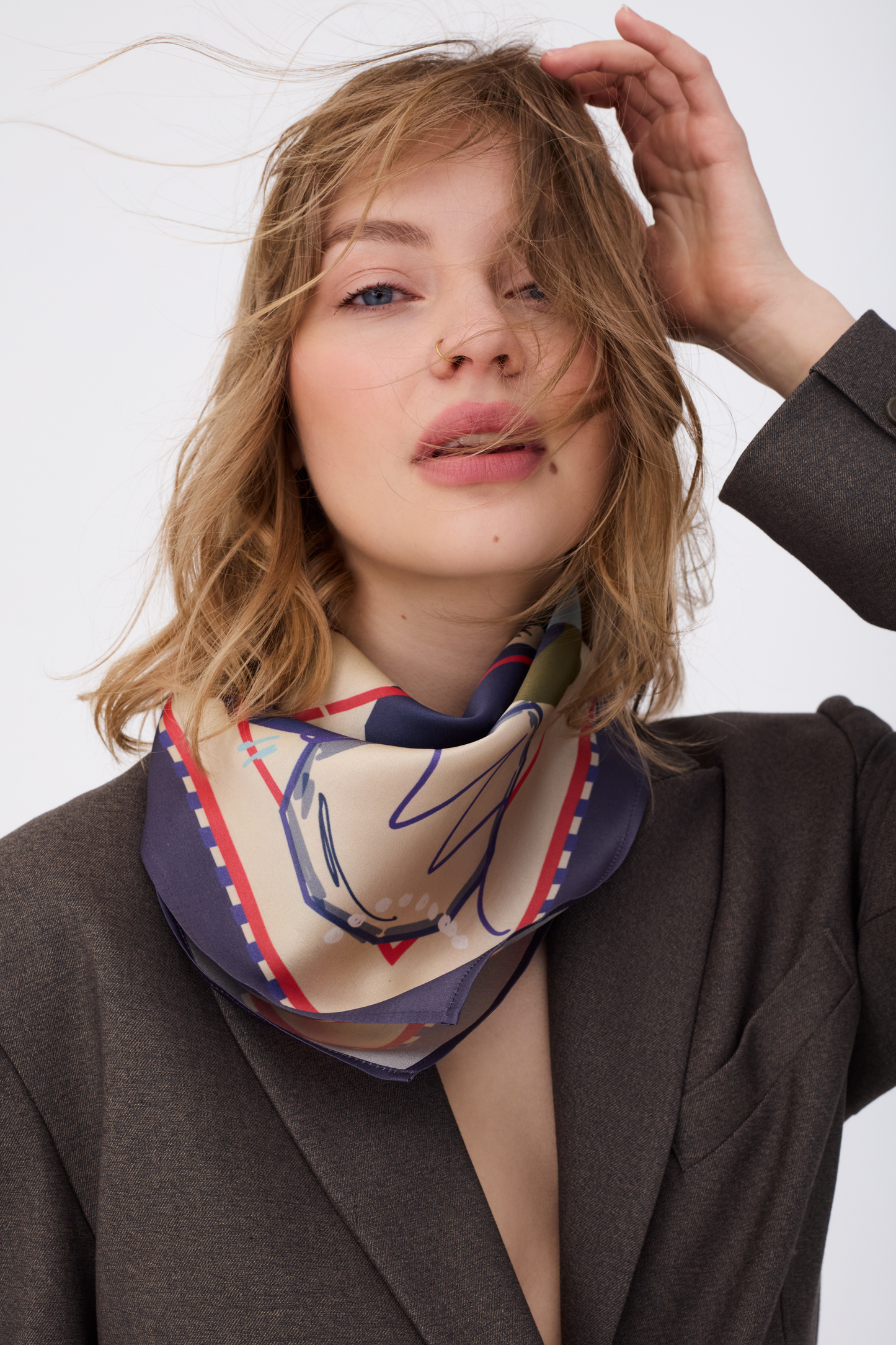 A close-up portrait of a woman with messy blonde hair, looking directly at the camera with her hand raised to her head. She is wearing the GUARDIAN Blue Graphite Silk Scarf tied loosely around her neck. The scarf's creamy center features a blue line-art terrier illustration, framed by a deep graphite blue border with a red and white dashed line. She is wearing a dark brown blazer against a bright white background
