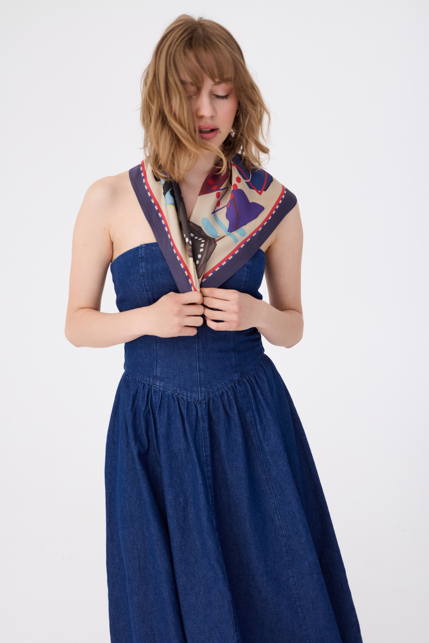 A woman with wavy blonde hair poses with her eyes closed, wearing the GUARDIAN Blue Graphite Silk Scarf draped neatly over her shoulders like a small shawl. The scarf's creamy base with its abstract terrier illustration in blue line art is prominent. She is wearing a strapless dark blue denim dress, creating a modern contrast against a plain white background.