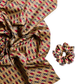 A luxury silk accessory set featuring a large scarf and a matching mid-size scrunchie with an intricate geometric diamond pattern in burgundy, gold, and black tones on a white background.