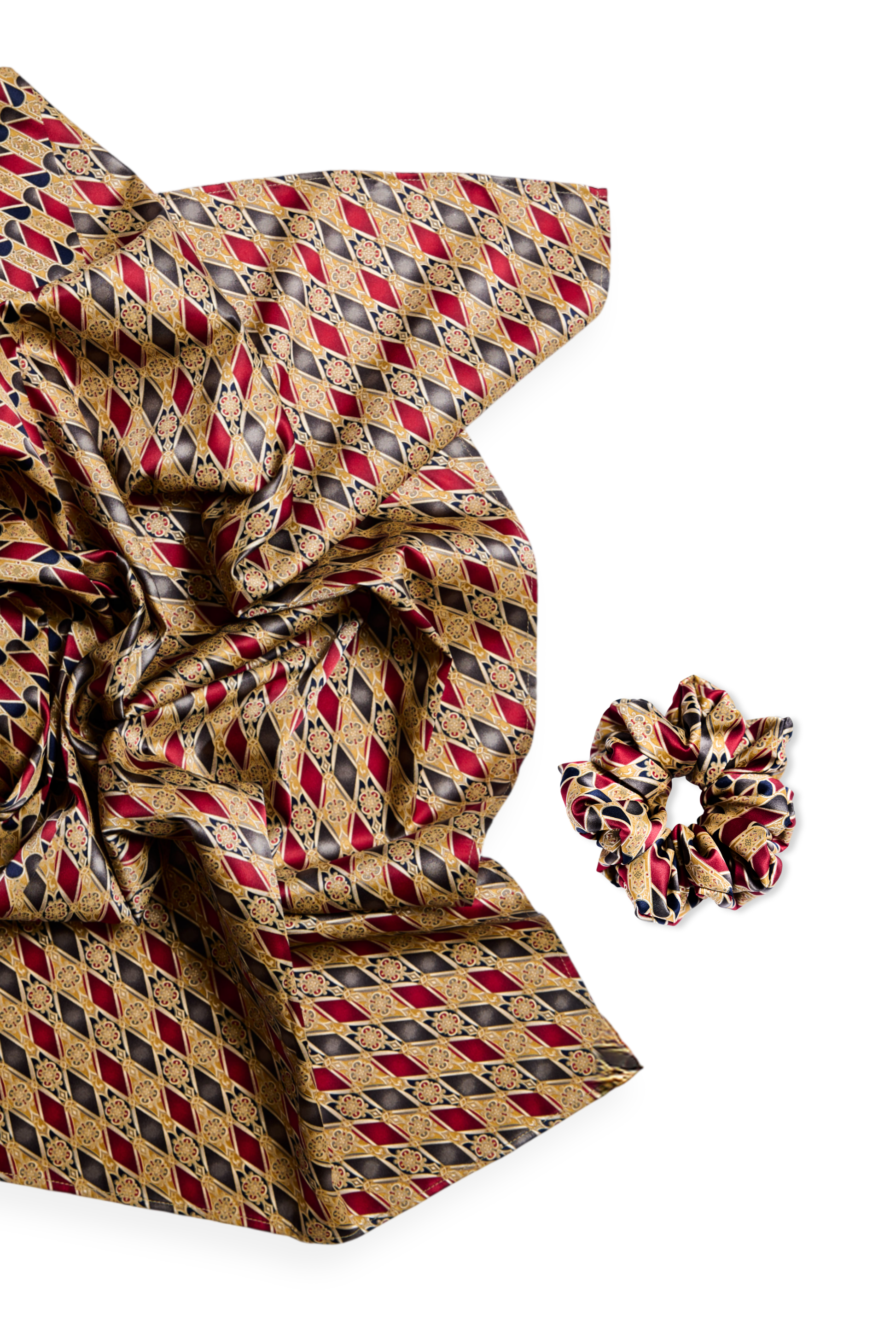 A luxury silk accessory set featuring a large scarf and a matching mid-size scrunchie with an intricate geometric diamond pattern in burgundy, gold, and black tones on a white background.