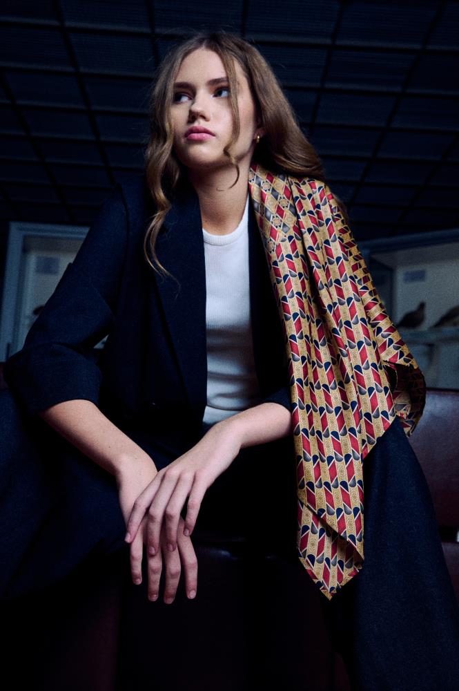 young woman with long wavy hair sits in a dramatically lit interior, looking to the side. She wears a dark blazer over a white t-shirt, with a wide silk scarf stylishly draped over her left shoulder and down her side. The scarf is decorated with a dense geometric diamond pattern dominated by red, gold, and black colors. Her hands are resting calmly in front, and the photo composition and outfit create a modern and elegant mood.