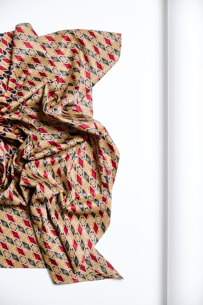 A crumpled, luxurious silk scarf laid out on the left side of a white background. The fabric is decorated with a dense, symmetrical geometric pattern consisting of red, gold, and black diamonds and stylized floral ornaments. The folds of the material and the soft surface sheen highlight the fabric's texture and rich, classic design, creating a dynamic and elegant composition.