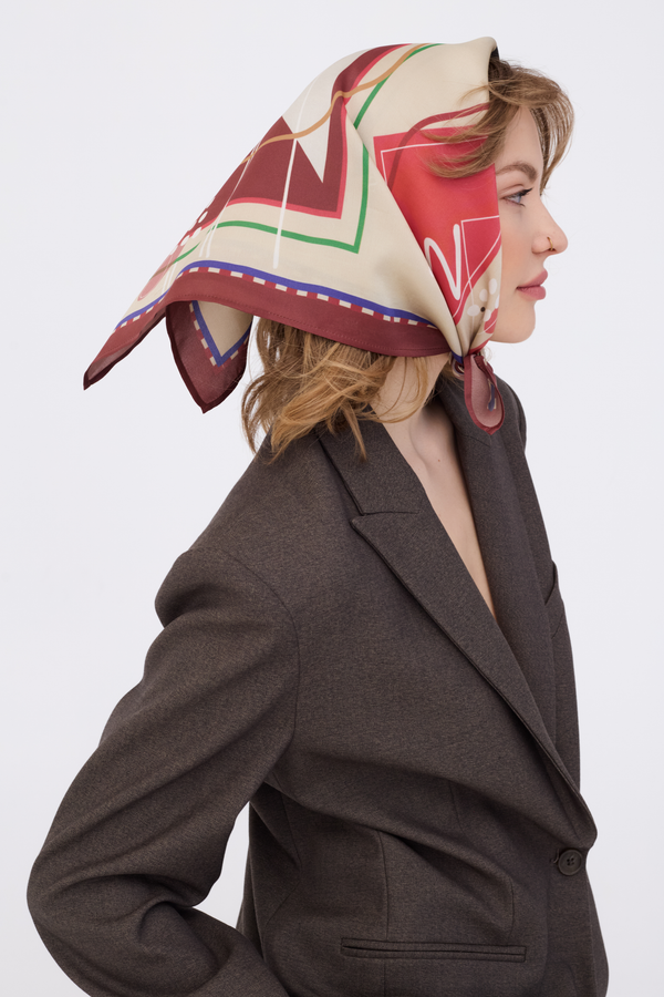 Side profile of a woman wearing the MUSE Chestnut Brown Silk Scarf tied elegantly over her head. The scarf features a sophisticated geometric pattern with chestnut brown, cream, and bold red accents. She is styled in a textured chocolate brown blazer against a clean white background.