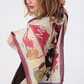 Detailed view of the MUSE Silk Scarf in Chestnut Brown, held open to reveal a modern abstract pattern of whimsical shapes and lines in terracotta, gold, and deep brown. The lightweight, lustrous silk fabric drapes naturally, highlighting its high-quality texture. The model is seen from the back and side, emphasizing the scarf's square dimensions.