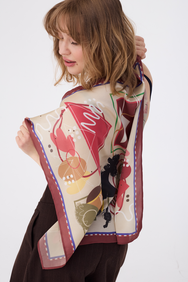 Detailed view of the MUSE Silk Scarf in Chestnut Brown, held open to reveal a modern abstract pattern of whimsical shapes and lines in terracotta, gold, and deep brown. The lightweight, lustrous silk fabric drapes naturally, highlighting its high-quality texture. The model is seen from the back and side, emphasizing the scarf's square dimensions.