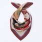 A flat lay view of the MUSE Chestnut Brown Silk Scarf, elegantly twisted to highlight its intricate abstract pattern. The design includes artistic sketches of figures and geometric shapes in earthy tones. The chestnut brown edges frame the multi-colored center, emphasizing the luxurious quality and modern artistic aesthetic of the silk accessory.
