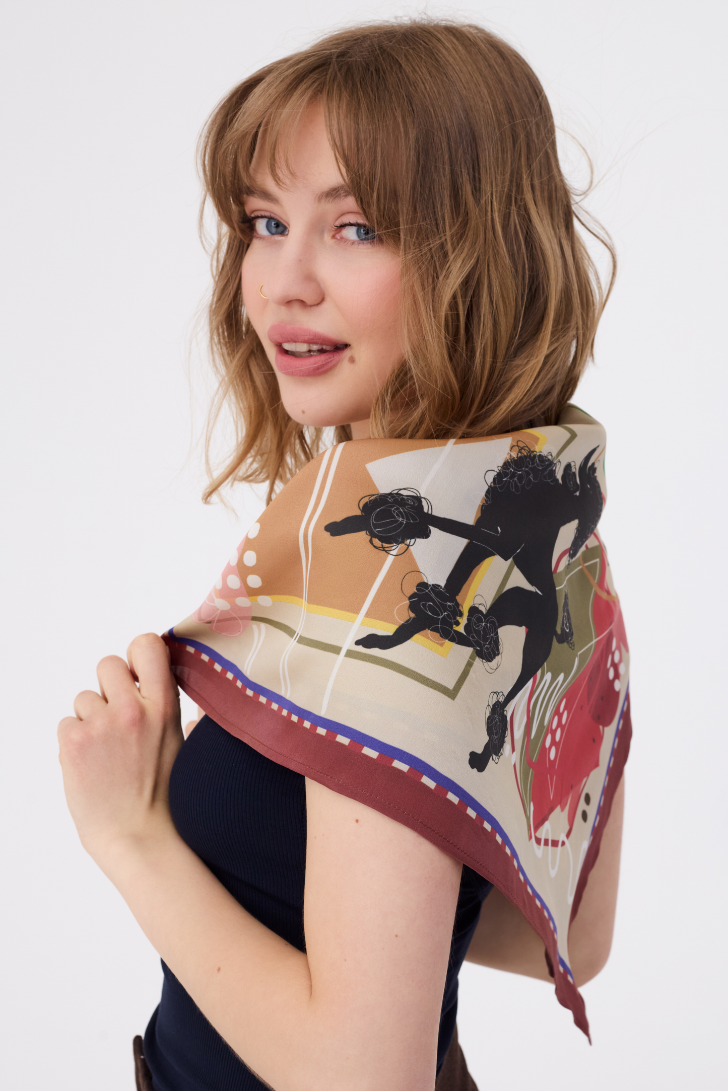 Detail of the MUSE Silk Scarf in Chestnut Brown, showcasing a whimsical black poodle silhouette as part of its modern abstract print. The scarf's creamy base is decorated with geometric lines and earthy tones, styled as a chic shoulder wrap. The image highlights the artistic storytelling and playful design of the accessory.