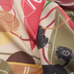 Macro shot of the MUSE Silk Scarf in Chestnut Brown, emphasizing the rich pigmentation of the fabric's dyes. The abstract pattern features terracotta red triangles, ochre circles, and dark line art. The way the fabric bunches shows its lightweight and fluid drape, perfect for luxury accessory lovers.