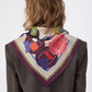 Back view of a woman with wavy blonde hair wearing the NOMAD Velvet Plum Silk Scarf draped over her shoulders in a triangular fold. The scarf features a deep plum purple border and an ivory center filled with abstract shapes in pink, orange, and blue. A stylized black sighthound illustration is visible near the neck. She is wearing a textured dark brown blazer against a white background