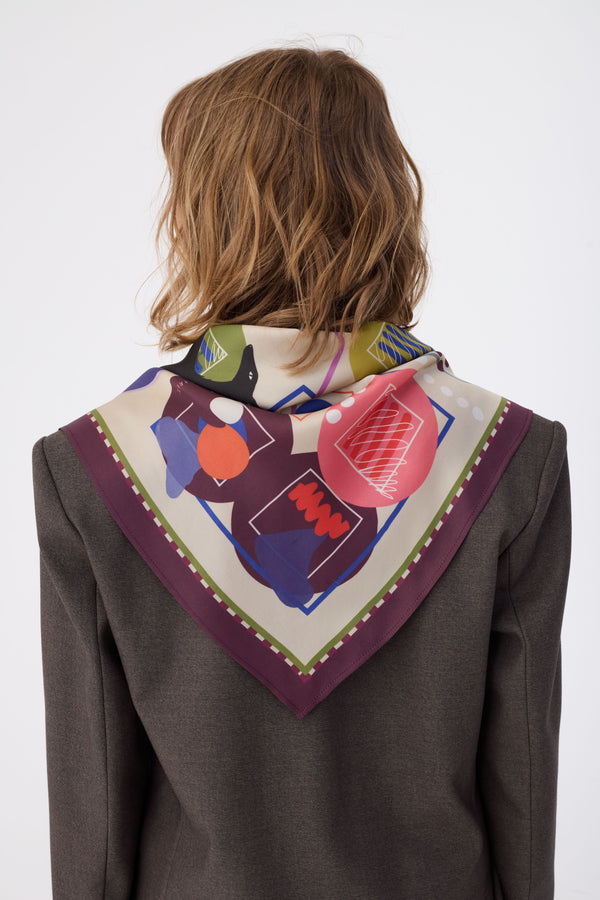 Back view of a woman with wavy blonde hair wearing the NOMAD Velvet Plum Silk Scarf draped over her shoulders in a triangular fold. The scarf features a deep plum purple border and an ivory center filled with abstract shapes in pink, orange, and blue. A stylized black sighthound illustration is visible near the neck. She is wearing a textured dark brown blazer against a white background