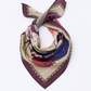 The NOMAD Velvet Plum Silk Scarf is shown folded in a neck-tie arrangement on a white background. The scarf features a wide, deep plum purple border with an intricate green and white dashed inner line. The center reveals a sophisticated abstract pattern with shades of magenta, navy blue, and sage green. A black sighthound silhouette is partially visible within the artistic folds of the smooth silk fabric.