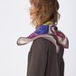 Close-up profile shot of the NOMAD Silk Scarf in Velvet Plum, styled dynamically. The movement of the wind catches the edges of the scarf, highlighting the light and fluid quality of the silk material. The intricate abstract design with the sighthound illustration and geometric shapes in autumn colors drapes around her neck, complemented by a tailored brown suit jacket.