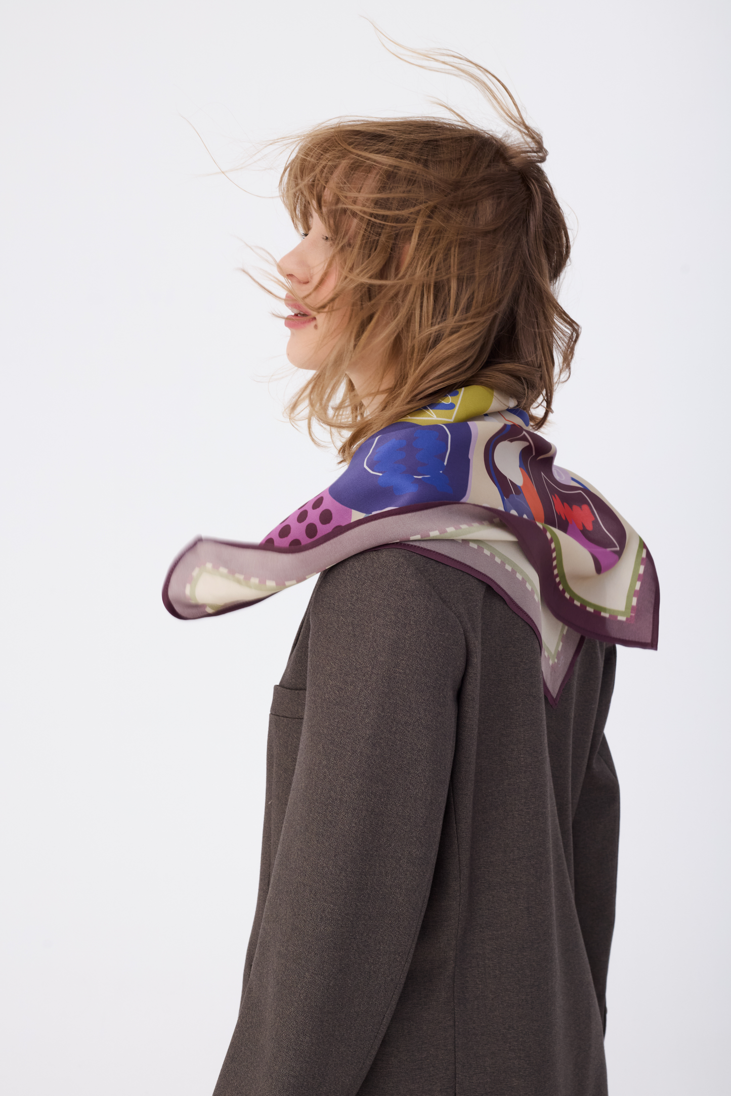 Close-up profile shot of the NOMAD Silk Scarf in Velvet Plum, styled dynamically. The movement of the wind catches the edges of the scarf, highlighting the light and fluid quality of the silk material. The intricate abstract design with the sighthound illustration and geometric shapes in autumn colors drapes around her neck, complemented by a tailored brown suit jacket.
