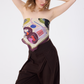 A woman with wavy blonde hair poses with her hands behind her head, wearing the NOMAD Velvet Plum Silk Scarf styled as a strapless tube top. The scarf is tied at the back, showcasing its central abstract pattern with magenta circles, navy blue shapes, and a black sighthound illustration. She is paired with high-waisted chocolate brown pinstripe trousers against a white background