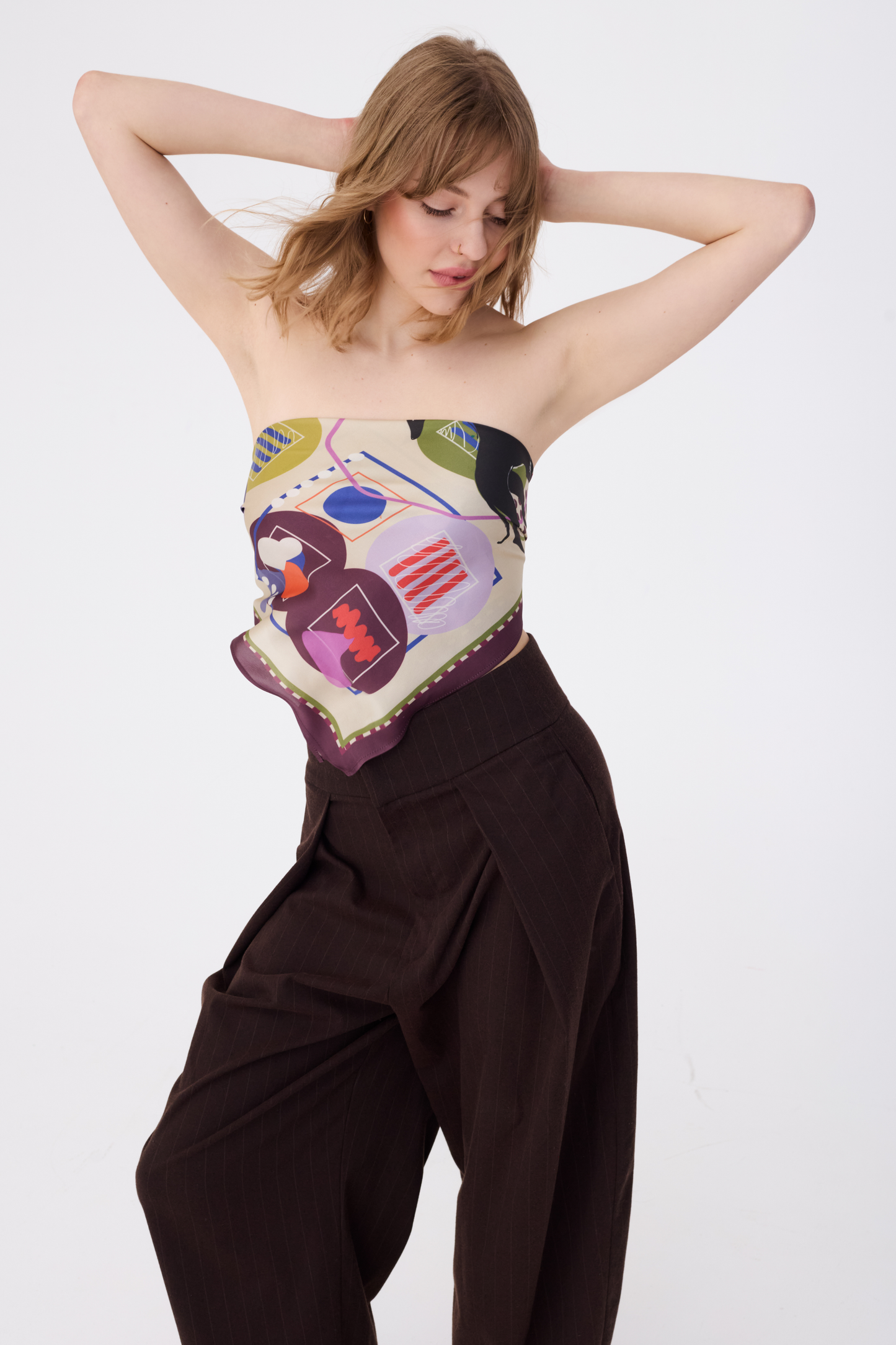 A woman with wavy blonde hair poses with her hands behind her head, wearing the NOMAD Velvet Plum Silk Scarf styled as a strapless tube top. The scarf is tied at the back, showcasing its central abstract pattern with magenta circles, navy blue shapes, and a black sighthound illustration. She is paired with high-waisted chocolate brown pinstripe trousers against a white background