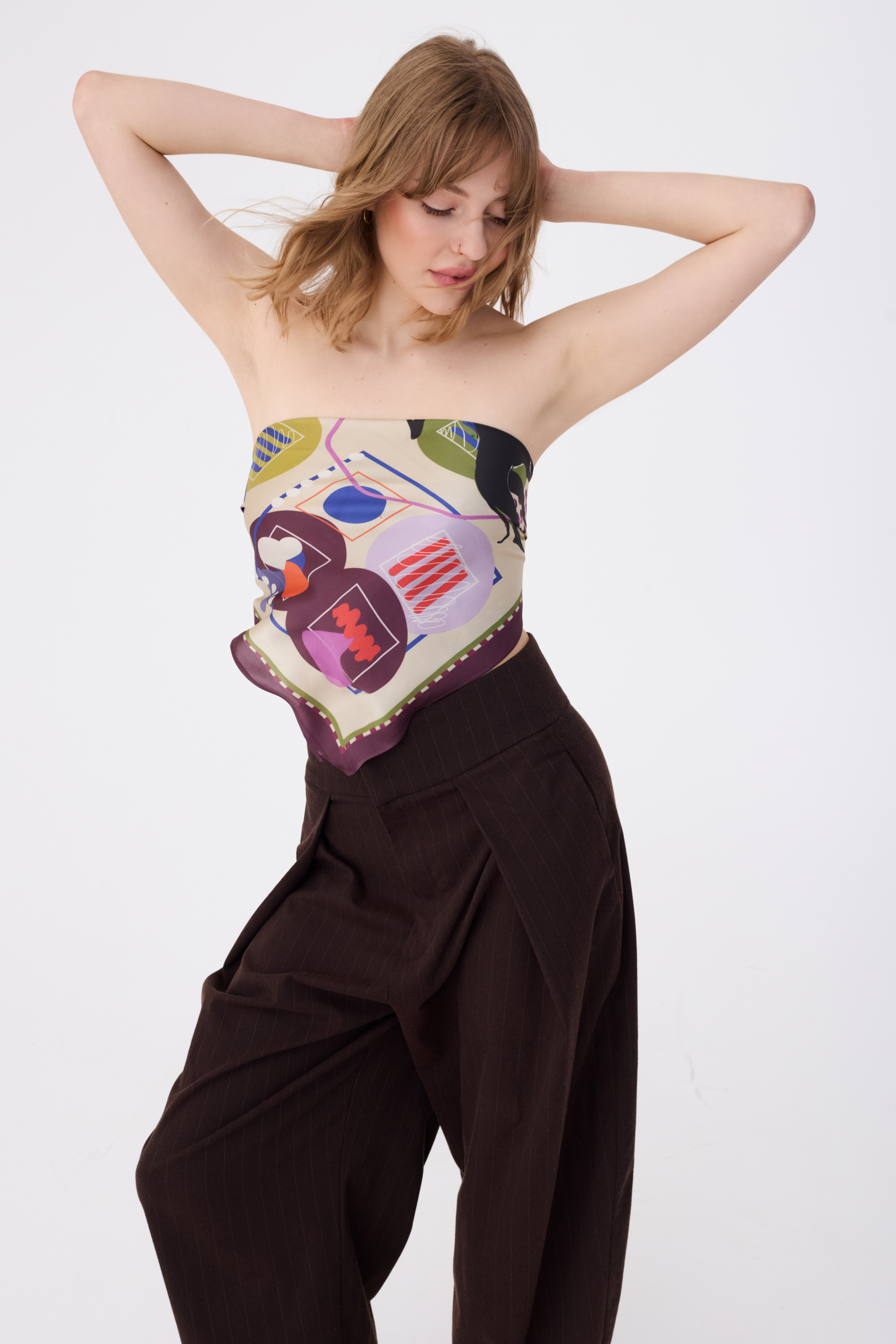 A woman with wavy blonde hair poses with her hands behind her head, wearing the NOMAD Velvet Plum Silk Scarf styled as a strapless tube top. The scarf is tied at the back, showcasing its central abstract pattern with magenta circles, navy blue shapes, and a black sighthound illustration. She is paired with high-waisted chocolate brown pinstripe trousers against a white background