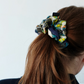 A back view of a girl with brown hair tied in a ponytail. Her hair is styled with a large, voluminous silk scrunchie featuring dark blue, green, and yellow patterns with tennis motifs. The background is light and neutral.