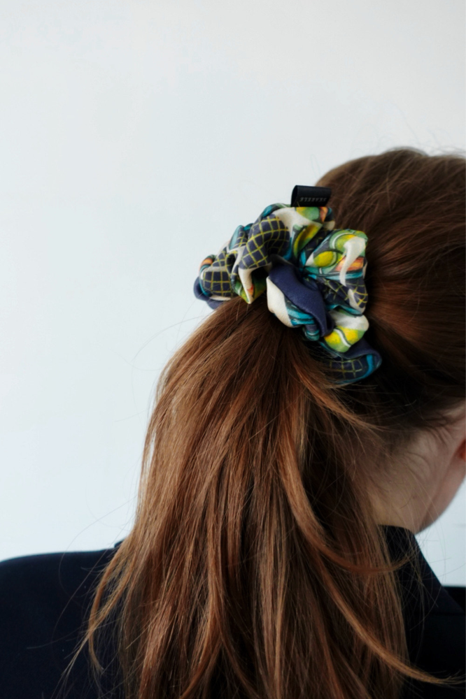 A back view of a girl with brown hair tied in a ponytail. Her hair is styled with a large, voluminous silk scrunchie featuring dark blue, green, and yellow patterns with tennis motifs. The background is light and neutral.