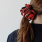 A back view of a young woman with long brown hair tied in a low ponytail. Her hair is styled with a large, voluminous red fabric scrunchie featuring black and cream patterns. She is wearing a dark blazer, and the photo is set against a light, neutral background.
