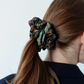 A back view of a girl with brown hair tied in a ponytail. Her hair is styled with a large, voluminous dark brown fabric scrunchie featuring green and orange botanical patterns. She is wearing a dark blazer, and the background is light and neutral.