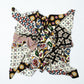 A square scarf laid out on a light background, with its center artistically crumpled into a swirl and the corners spread out. The scarf features a vibrant, eclectic pattern rich in geometric shapes, floral motifs, and mosaic details. The color palette is dominated by white, black, yellow, burgundy, and green.