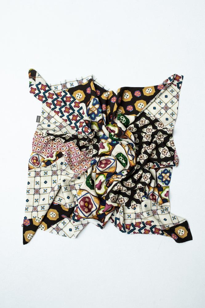 A square scarf laid out on a light background, with its center artistically crumpled into a swirl and the corners spread out. The scarf features a vibrant, eclectic pattern rich in geometric shapes, floral motifs, and mosaic details. The color palette is dominated by white, black, yellow, burgundy, and green.