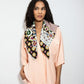 A woman with dark curly hair poses against a light background, wearing a loose peach-colored shirt and trousers set. A large square scarf with a vibrant, eclectic pattern—combining black, white, burgundy, and green motifs—is draped over her shoulders. She is looking to the side with one hand in her pocket, while the scarf ends hang freely in front.