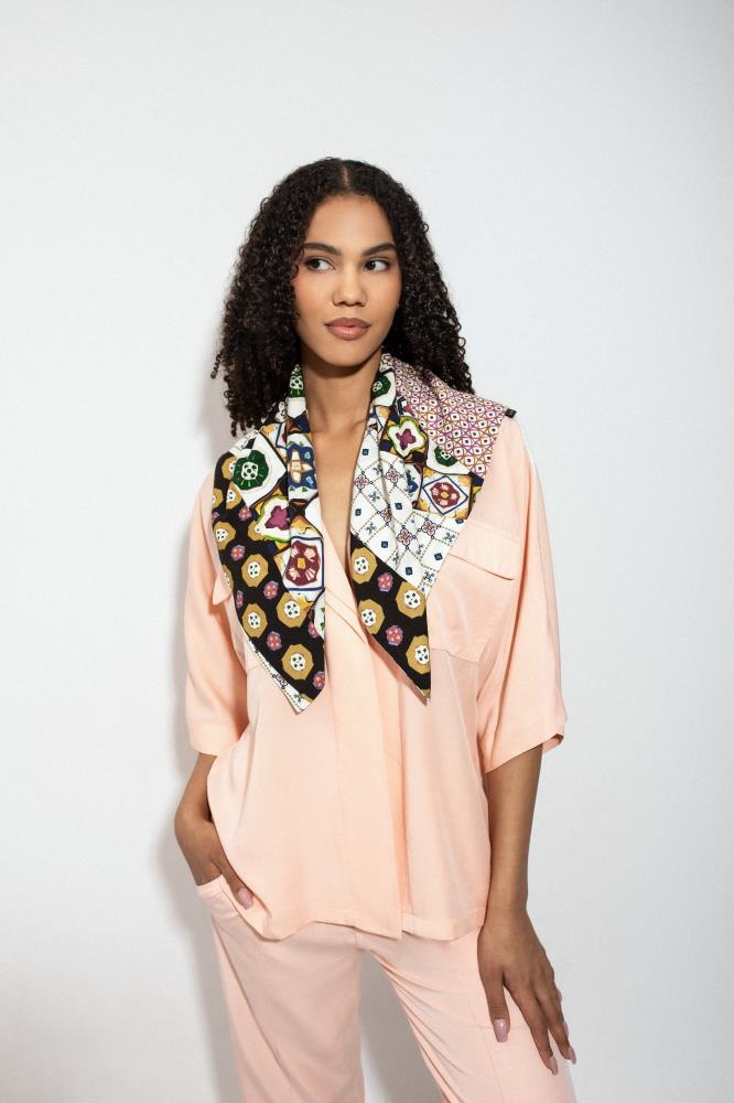 A woman with dark curly hair poses against a light background, wearing a loose peach-colored shirt and trousers set. A large square scarf with a vibrant, eclectic pattern—combining black, white, burgundy, and green motifs—is draped over her shoulders. She is looking to the side with one hand in her pocket, while the scarf ends hang freely in front.