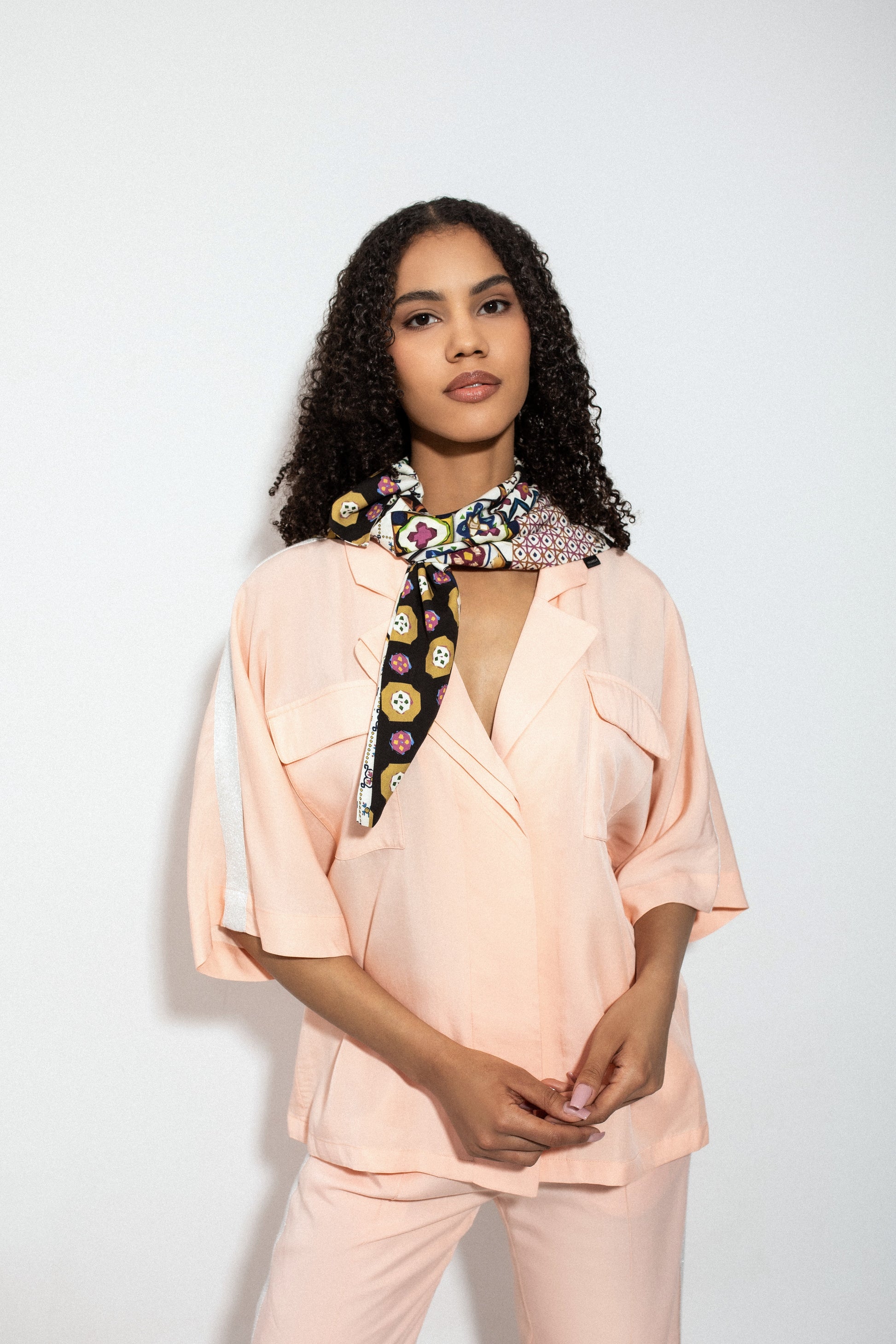 Woman wearing a peach blouse and pants with a colorful scarf around her neck against a white background