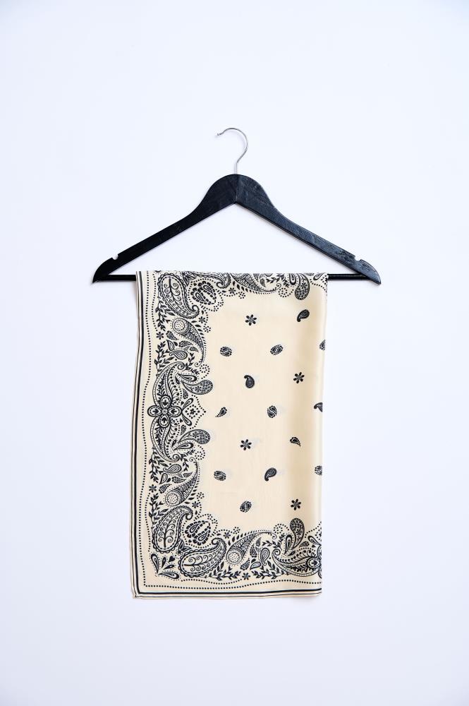 A square, light cream-colored scarf folded and hanging on a black wooden hanger against a white background. The scarf is decorated with a classic black paisley pattern, which is most dense along the edges, with smaller motifs and flowers repeating in the center. The composition is minimalist and tidy, highlighting the accessory's pattern and the smoothness of the fabric.