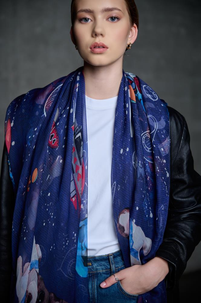 A portrait of a woman posing with a long, dark blue scarf featuring playful space-themed illustrations like planets and rockets. She is wearing a white t-shirt, a black leather jacket, and jeans. The model looks directly at the camera, and the dark, neutral background highlights the blue tones and colorful illustrations of the scarf, creating a modern and stylish look.